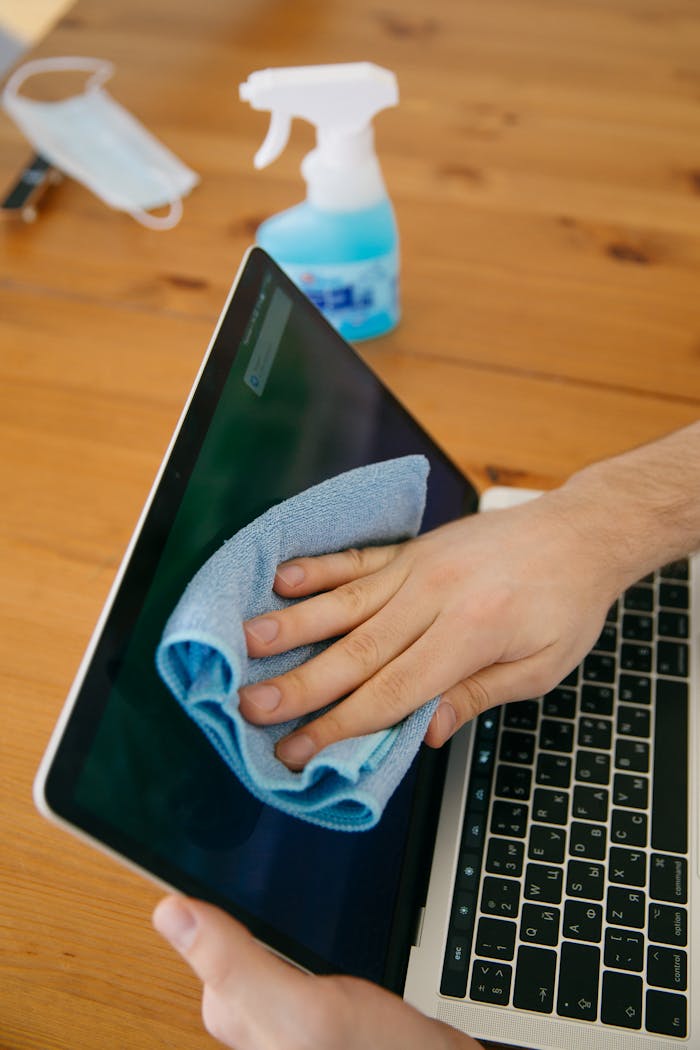 A person wipes a laptop screen using a blue cloth and spray bottle on a wooden table for hygiene.