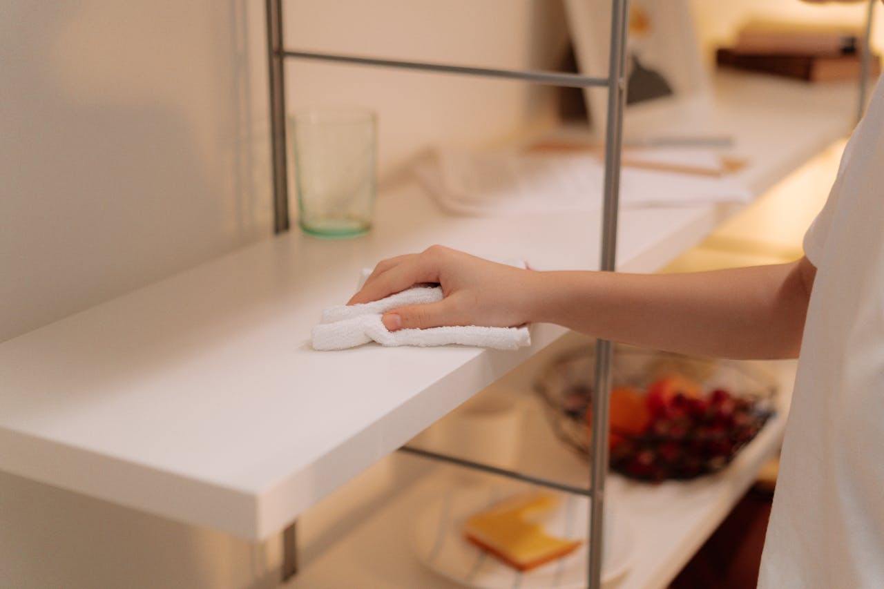 A person cleans a white shelf with a cloth, focusing on home maintenance.